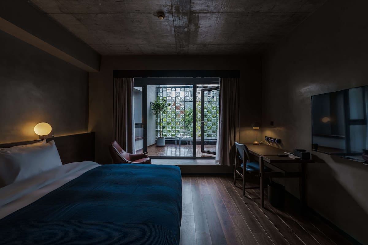 A hotel room with a bed, desk, chair, and a view of the outside through a glass door.