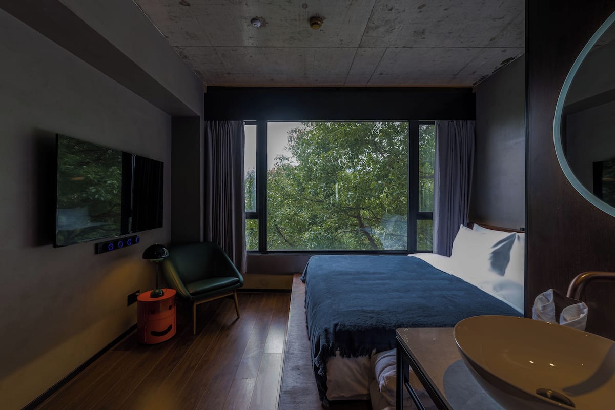 A modern hotel room with a large bed, a TV mounted on the wall, a desk with a sink, and a view of trees through the window.