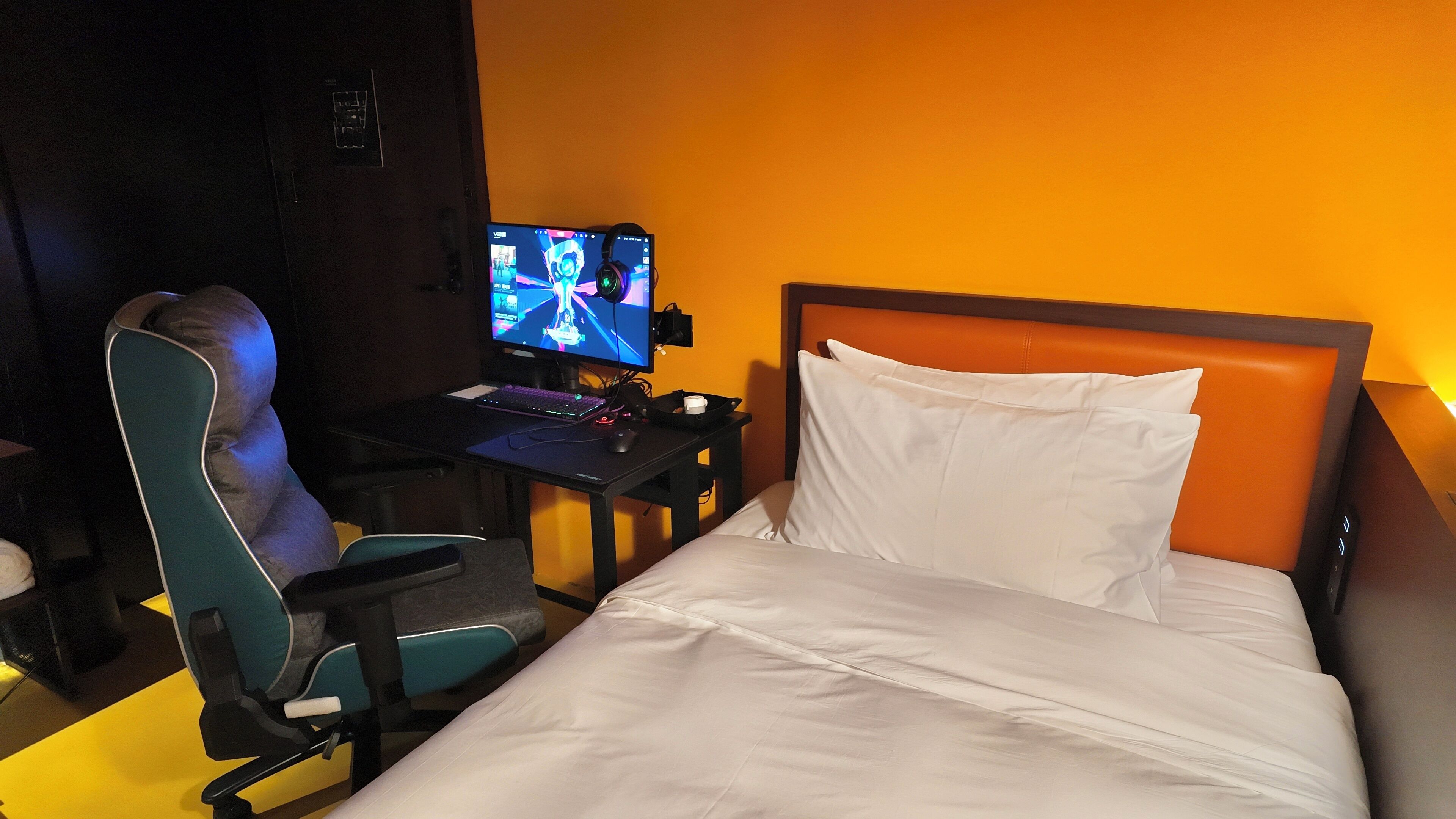 A hotel room with a bed, a desk with a computer and headphones, and a gaming chair.
