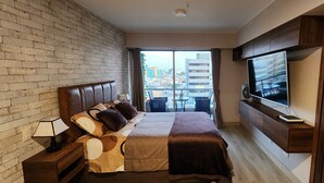 Una habitación de hotel moderna con pared de ladrillo, una cama grande con cabecera, un televisor de pantalla plana montado en la pared y un balcón con vista a la ciudad.