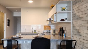 Una cocina moderna con encimera de granito, horno empotrado y cafetera. Hay dos sillas en la encimera y un estante pequeño con objetos decorativos.