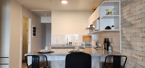 Een moderne keuken met een granieten aanrecht, een inbouwoven en een koffieapparaat. Er staan twee stoelen bij het aanrecht en een klein schap met decoratieve objecten.