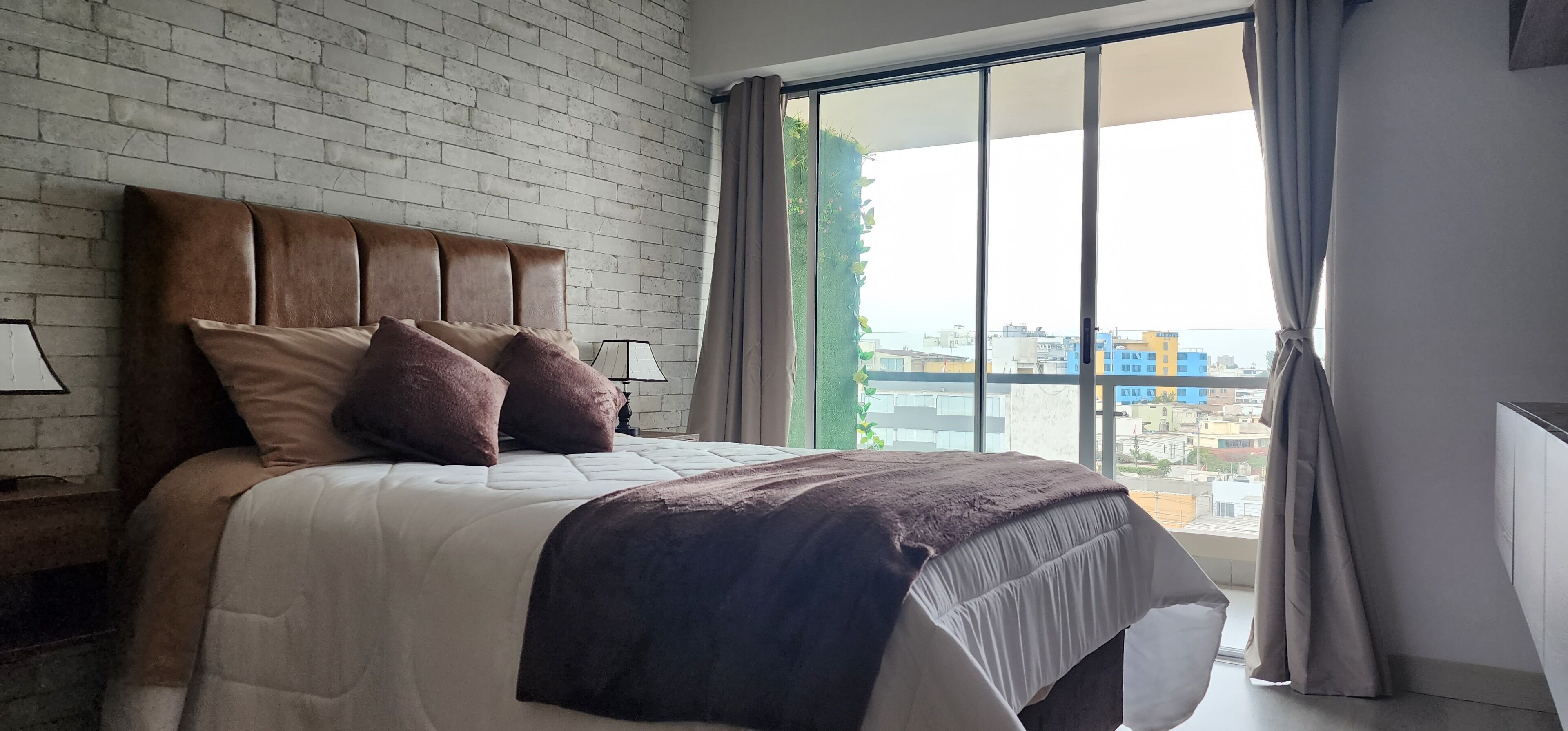 A modern bedroom with a brick wall, a large bed with a headboard, and a view of buildings through a large window.