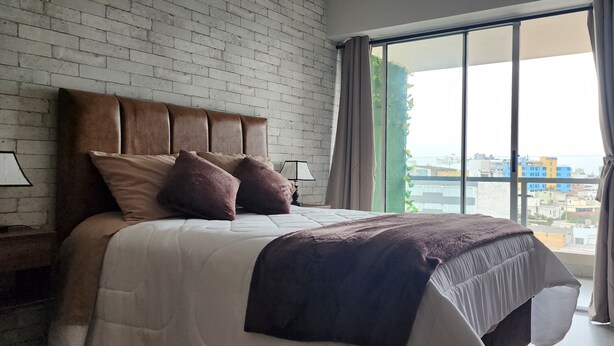 Un dormitorio moderno con pared de ladrillo, una cama grande con cabecera y vistas a edificios a través de un ventanal.