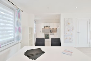 Una cocina moderna con armarios blancos, una estufa y una cafetera. Hay una mesa de comedor con sillas y una ventana con persianas.