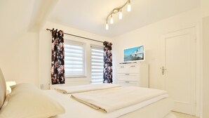 Ein Schlafzimmer mit einem Bett, zwei Fenstern mit Jalousien, einer weißen Kommode, einem an der Wand montierten Fernseher und einer Tür.