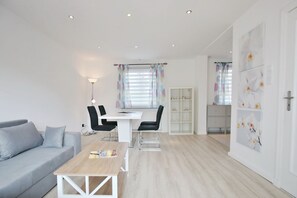 Una sala moderna con un sofá gris, una mesa de centro de madera y una mesa de comedor blanca con sillas. Se observa una estantería y una puerta con papel tapiz floral.