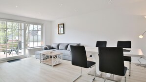 Ein modernes Wohnzimmer mit einer Couch, einem Couchtisch und Sesseln. Ein großes Fenster lässt natürliches Licht herein und bietet einen Ausblick ins Freie.