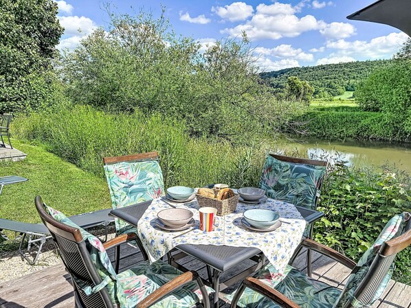 Un espace de restauration en plein air avec une table dressée pour quatre personnes, entouré de chaises à motifs floraux, offrant une vue sur une rivière et une végétation luxuriante.