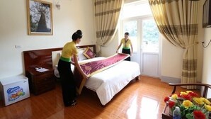 A hotel room with a wooden floor, a bed with a white and purple bedspread, a nightstand, a lamp, and a window with curtains.