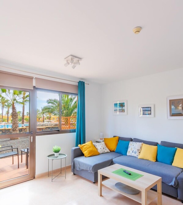Un salon moderne avec un canapé gris, des coussins colorés, une petite table et un balcon donnant sur la piscine.