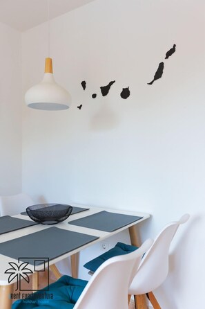 Un coin repas avec une table blanche, des sets de table noirs, un saladier en métal ajouré et des chaises blanches.