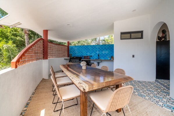 A dining area with a long wooden table, six chairs, and a kitchen in the background. - Casa Stellor - Modern, Spacious, Salt Pool, Walk to town center (Sayulita)
