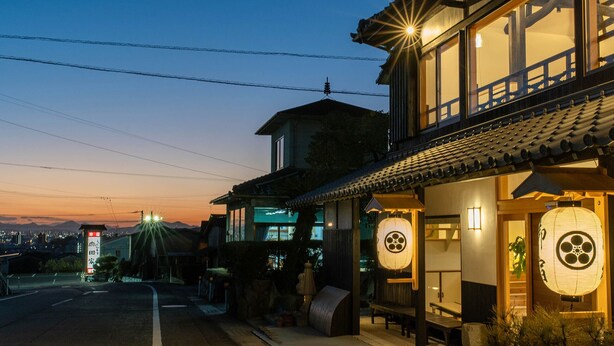 Un edificio japonés tradicional con fachada de madera y tejado de tejas, que cuenta con amplias ventanas y un farolillo exterior.