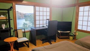 Una habitación con escritorio, silla y cama, una ventana con vista a un lago, y una estantería con una cesta y flores.