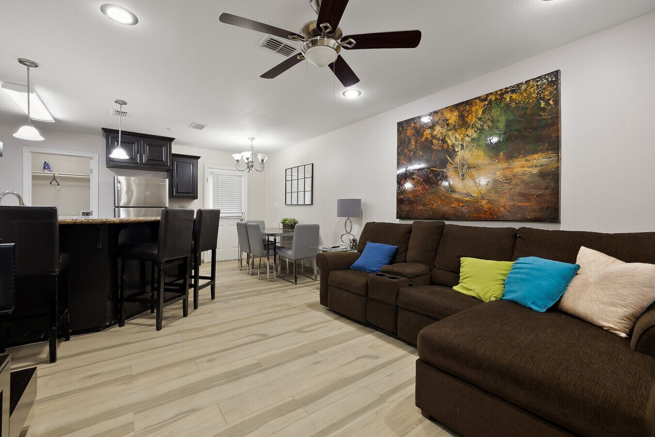 A modern living room with a brown sofa, dining area, kitchen, and ceiling fan.