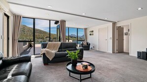 En rummelig stue med stor skydedør til en balkon, en sort lædersofa, et sofabord med en plante og udsigt til et bjerglandskab.
