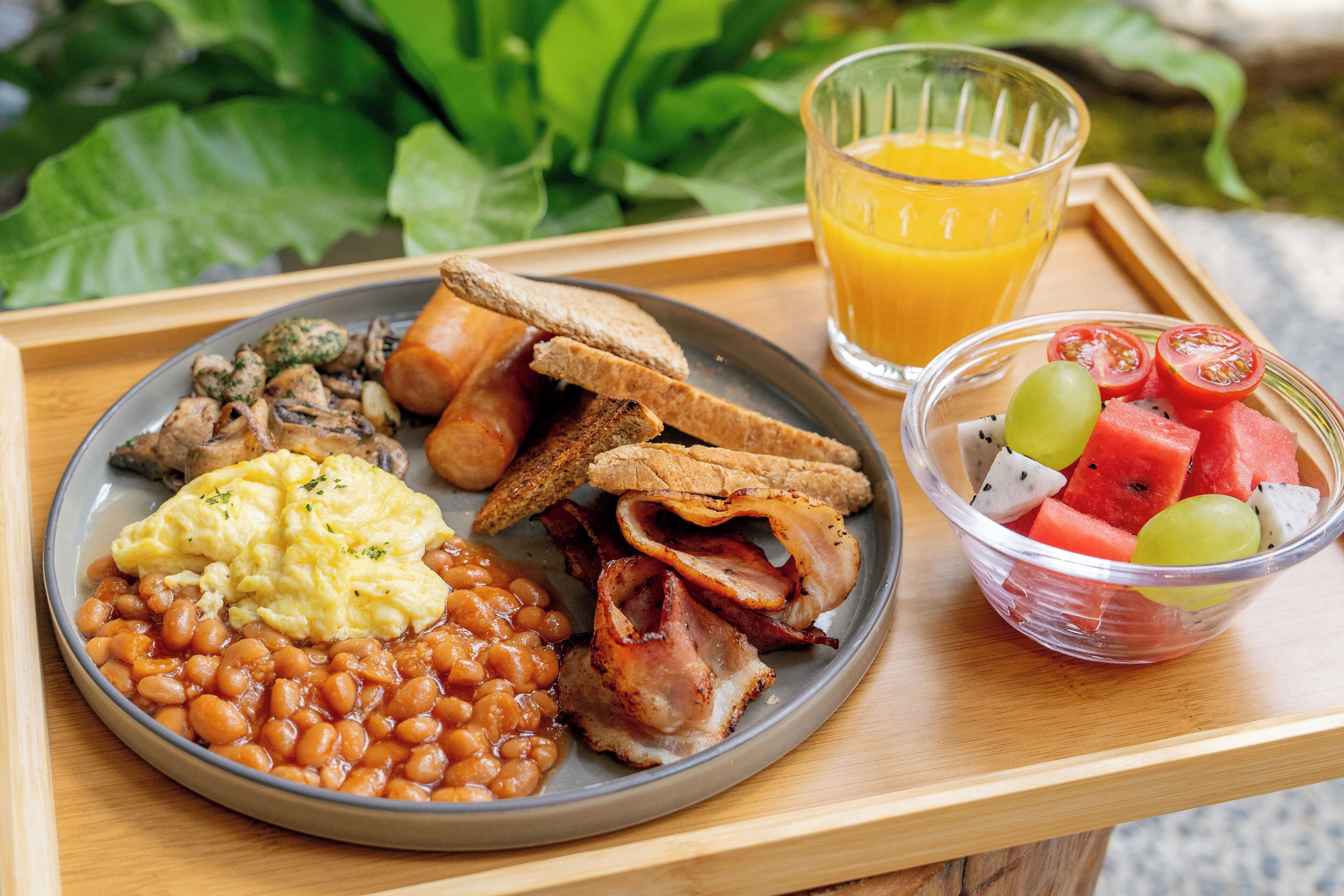 En frukostbricka med ägg, bönor, bacon, rostat bröd och en skål med frukt.