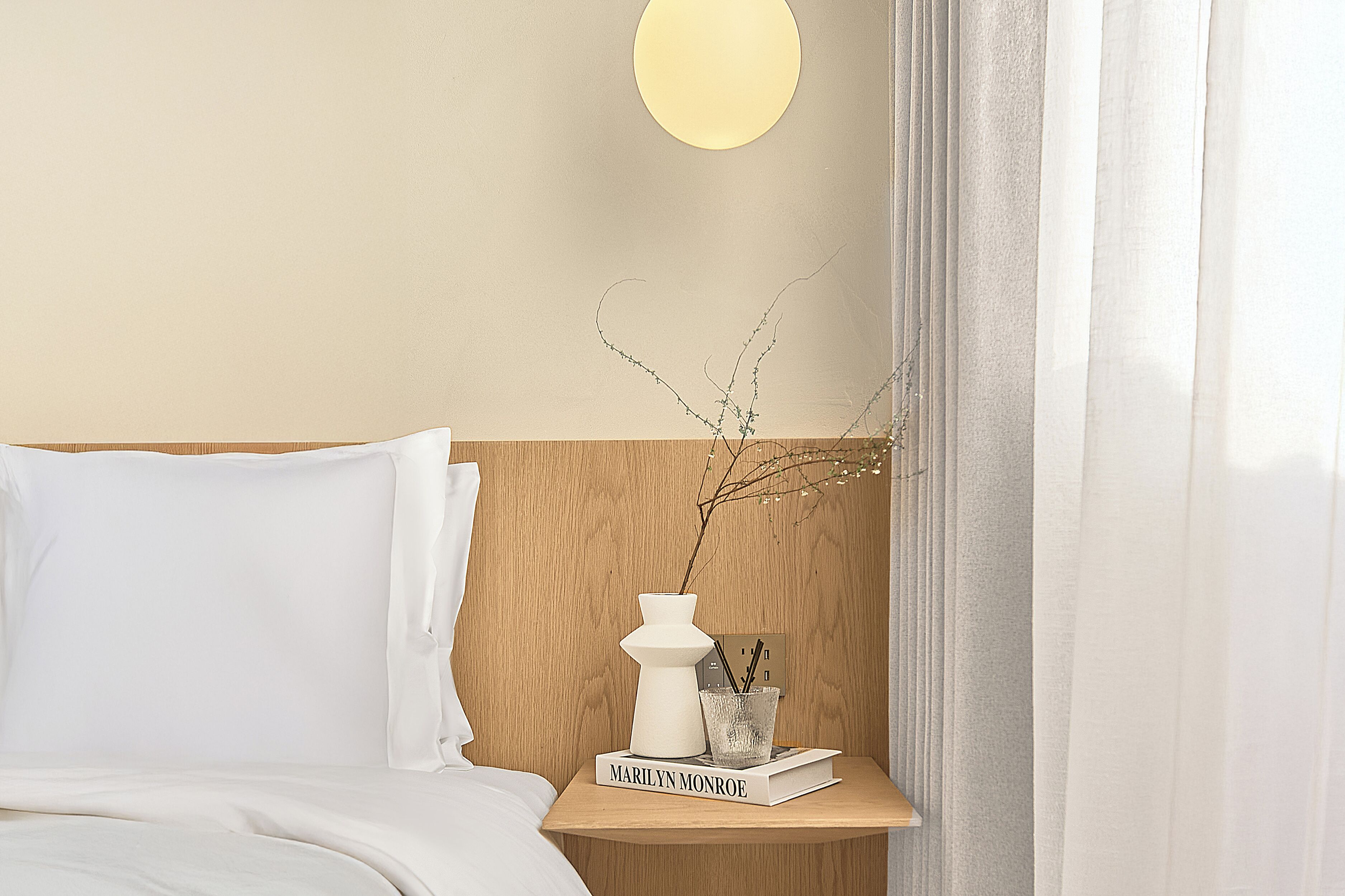 Una cama bien hecha con ropa blanca, una mesita de noche con un libro titulado 'Marilyn Monroe', un jarrón blanco con ramas y una lámpara de pared.