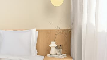Una cama bien hecha con ropa blanca, una mesita de noche con un libro titulado 'Marilyn Monroe', un jarrón blanco con ramas y una lámpara de pared.