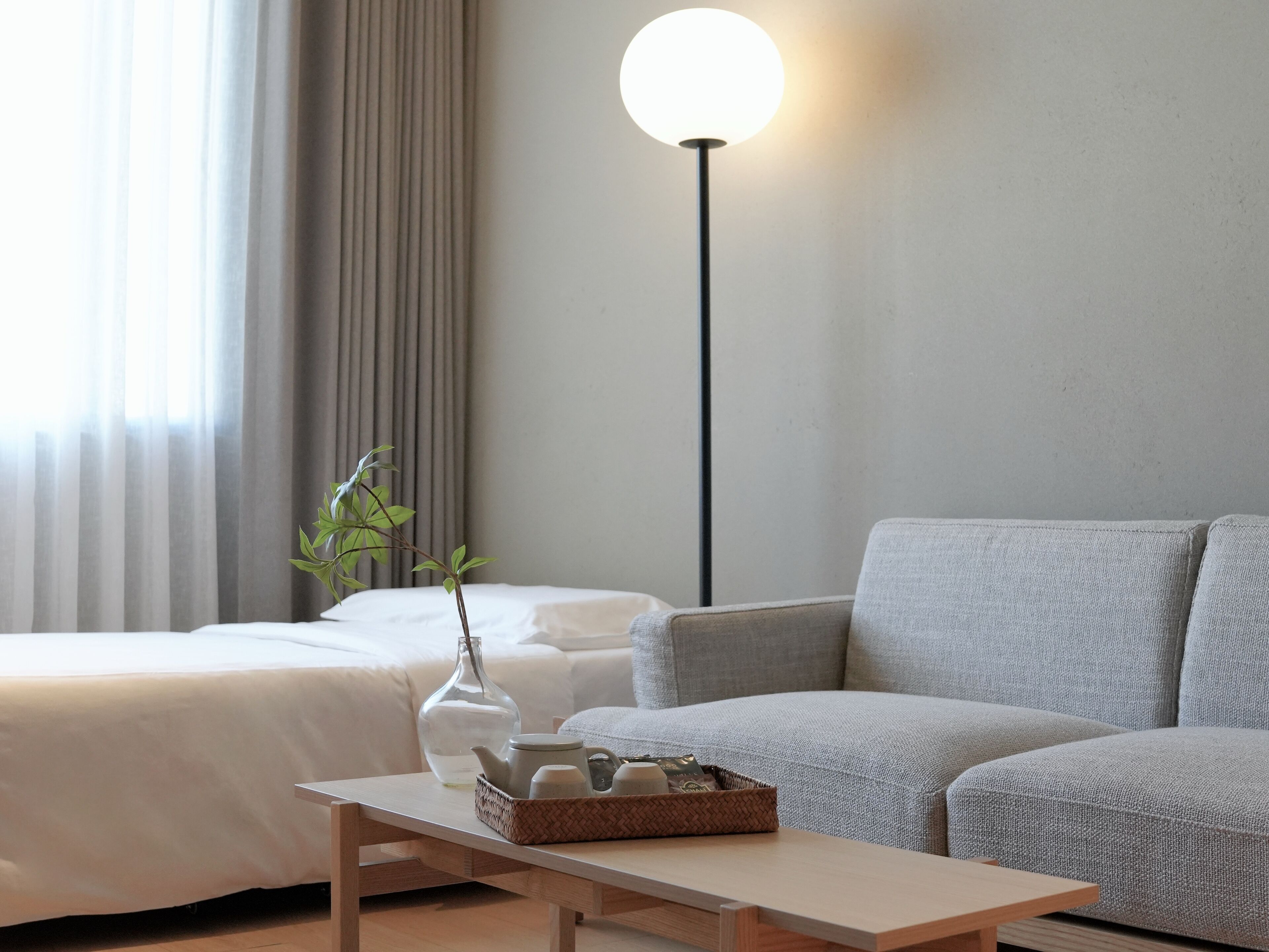 A modern hotel room with a bed, a sofa, a bedside table with a vase and a tray of toiletries, and a floor lamp.