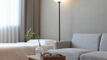 A modern hotel room with a bed, a sofa, a bedside table with a vase and a tray of toiletries, and a floor lamp.