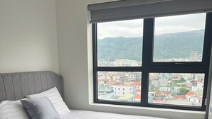 A bedroom with a bed, a window with a view of a town, and a grey headboard.