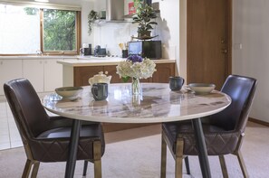 A dining area with a round table, two chairs, a vase with flowers, and kitchen appliances in the background. - Botanica Retreat - Village Spa & Stay (Olinda)