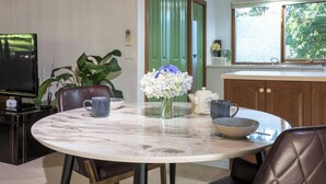 A dining area with a marble table, chairs, a vase with flowers, a television, and a kitchen in the background. - Botanica Retreat - Village Spa & Stay (Olinda)