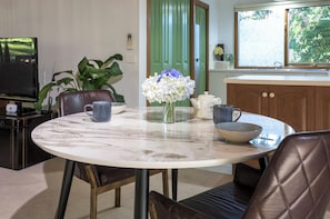A dining area with a marble table, chairs, a vase with flowers, a television, and a kitchen in the background. - Botanica Retreat - Village Spa & Stay (Olinda)