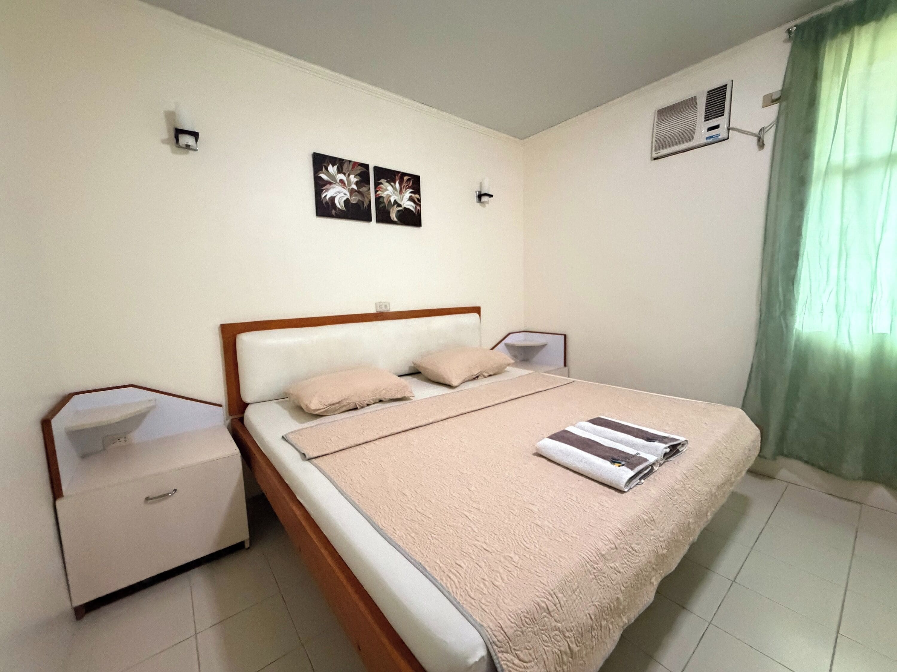 Un dormitorio ordenado con cama, mesitas de noche, aire acondicionado instalado en la pared y una ventana con cortinas.
