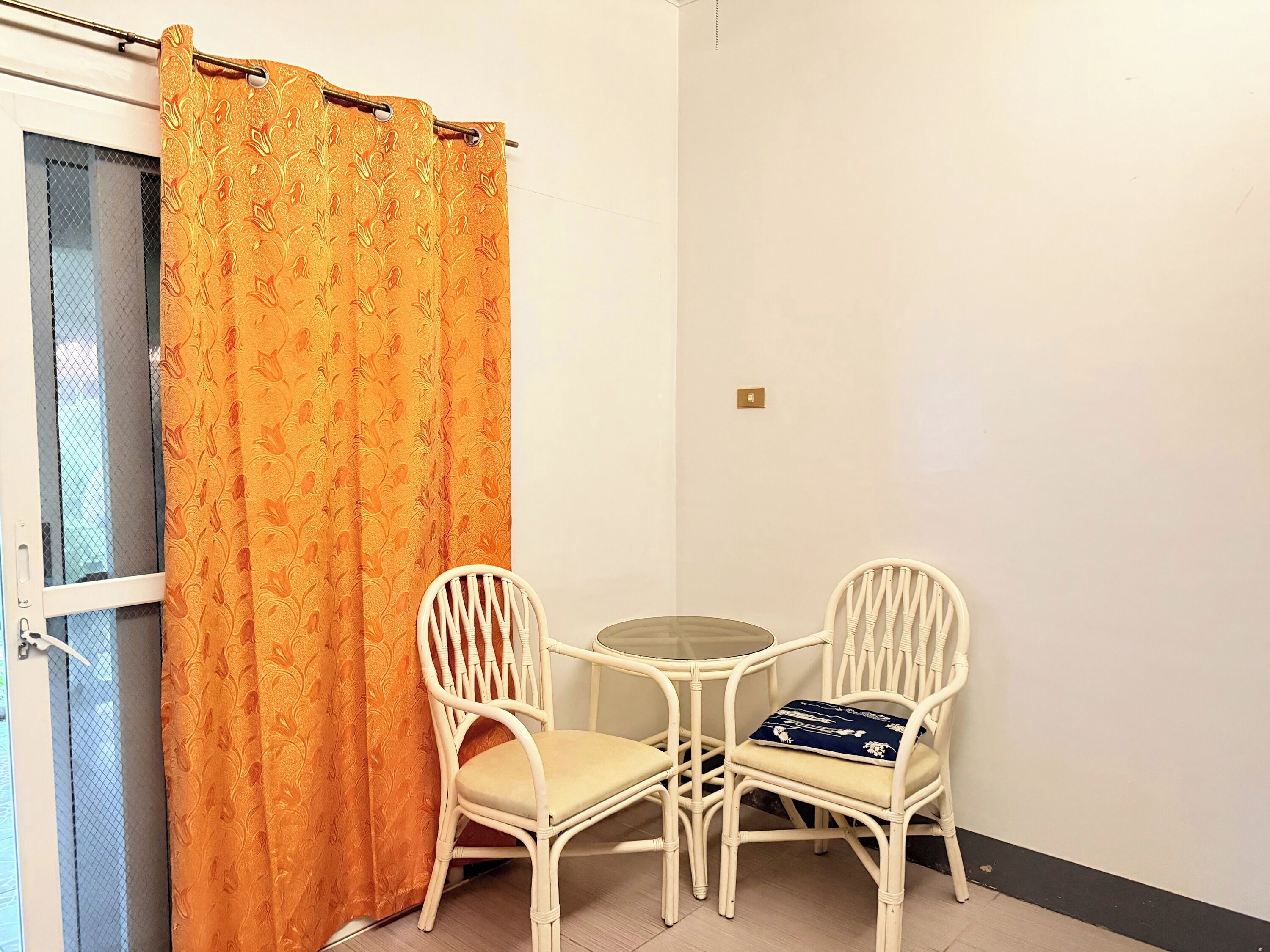 A small room with two white wicker chairs, a round table, and an orange patterned curtain.