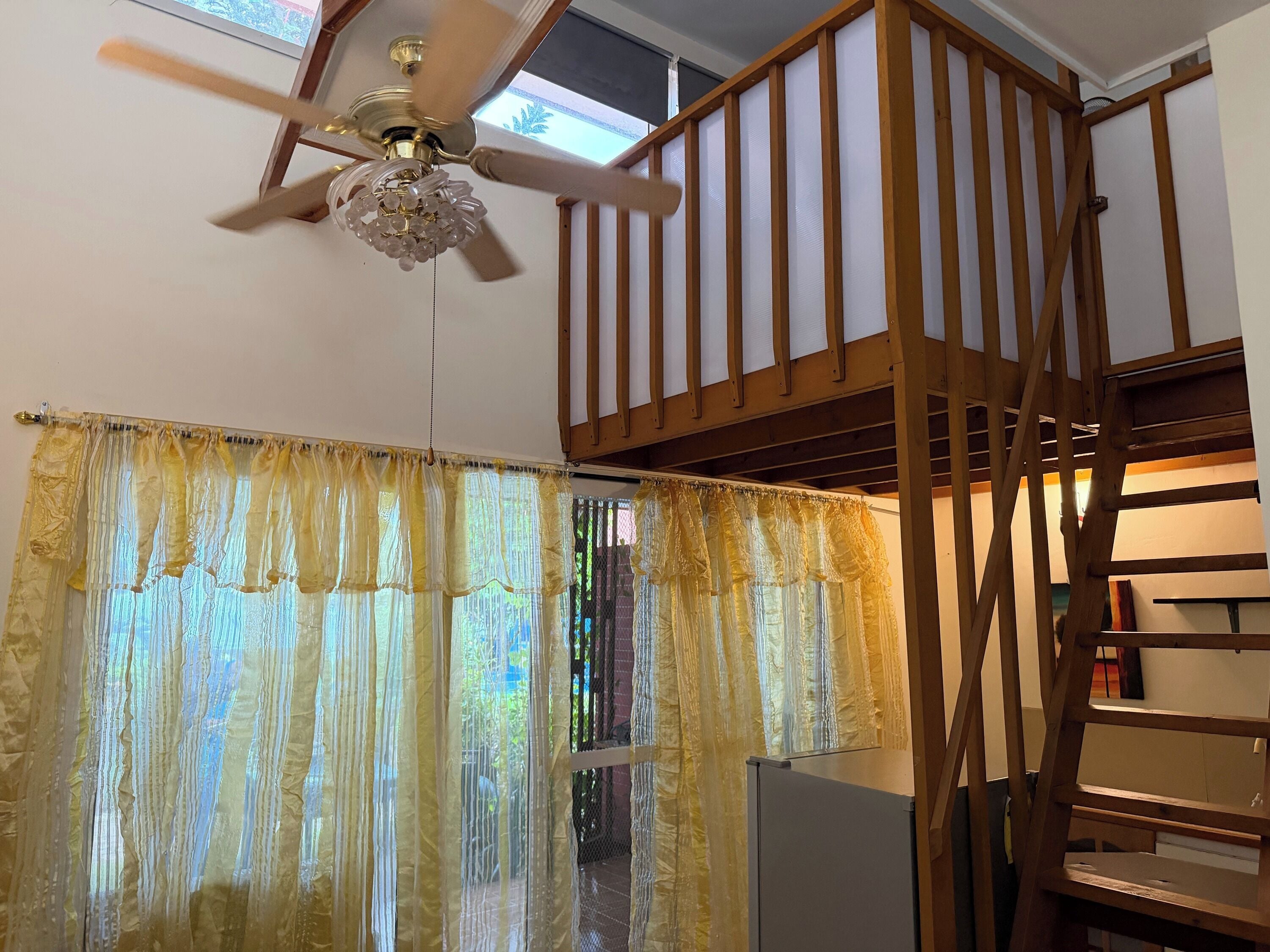 Una habitación con ventilador de techo, una escalera de madera y una ventana con cortinas traslúcidas.