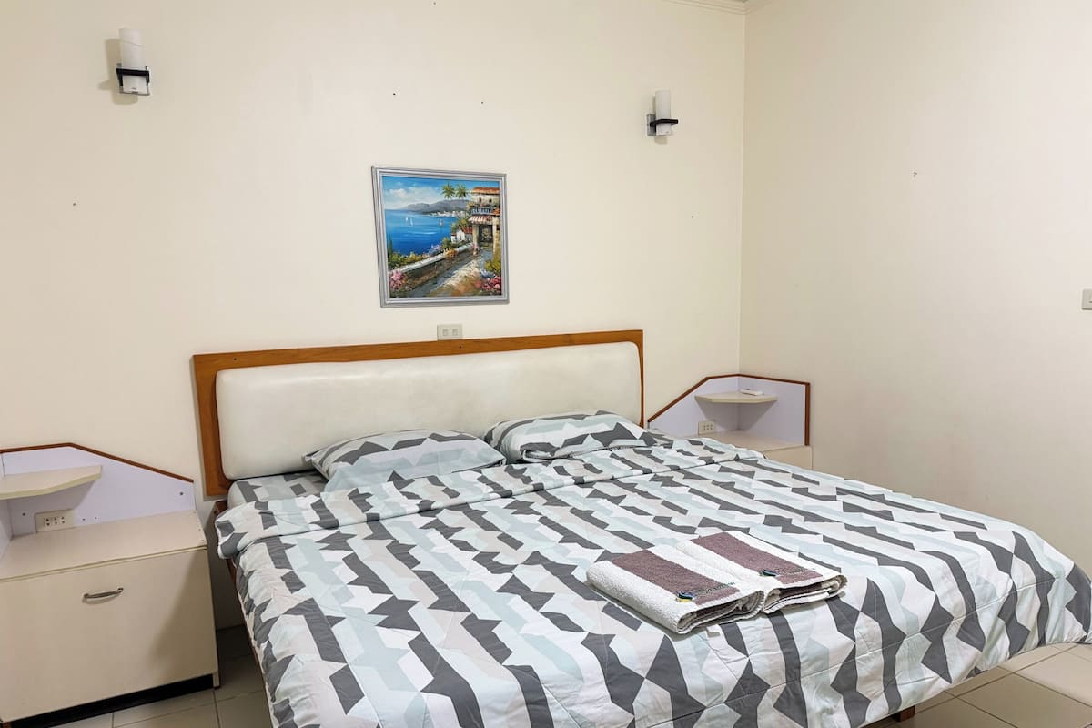 A neatly made bed with a geometric-patterned bedspread, a wooden headboard, a bedside table with a lamp, a small shelf, and a wall-mounted picture of a coastal scene.
