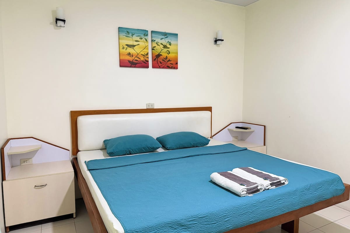 A neatly made bed with a blue bedspread and pillows, a bedside table with a remote control, and two wall-mounted light fixtures.