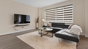 Een moderne woonkamer met een hoekbank, een flatscreen televisie aan de muur, een salontafel en een groot raam met zonwering.