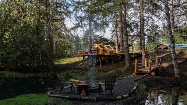 Een vakantiehuis op palen, omringd door bos, met een vuurkuil en stoelen op de voorgrond.