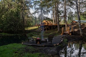 A cabin on stilts surrounded by forest, with a fire pit and chairs in the foreground.