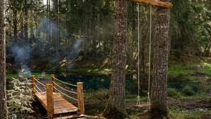 Een houten brug over een beek in een bebost gebied, met stoelen en een schommel.