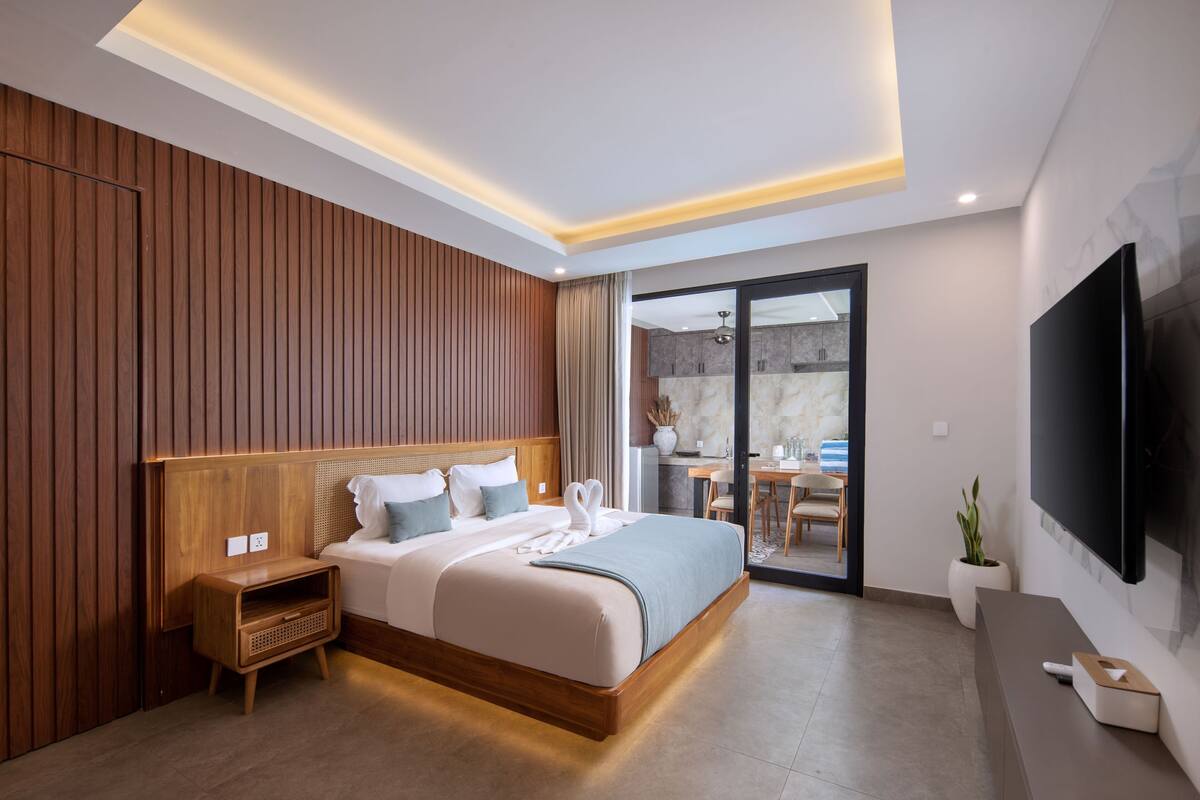 A modern bedroom with a wooden headboard, a large bed with white linens, a bedside table, a flat-screen TV, and a potted plant.