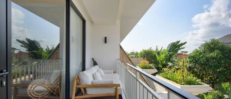 A balcony with a white sofa and a wooden chair, a glass door, and a view of greenery and buildings.