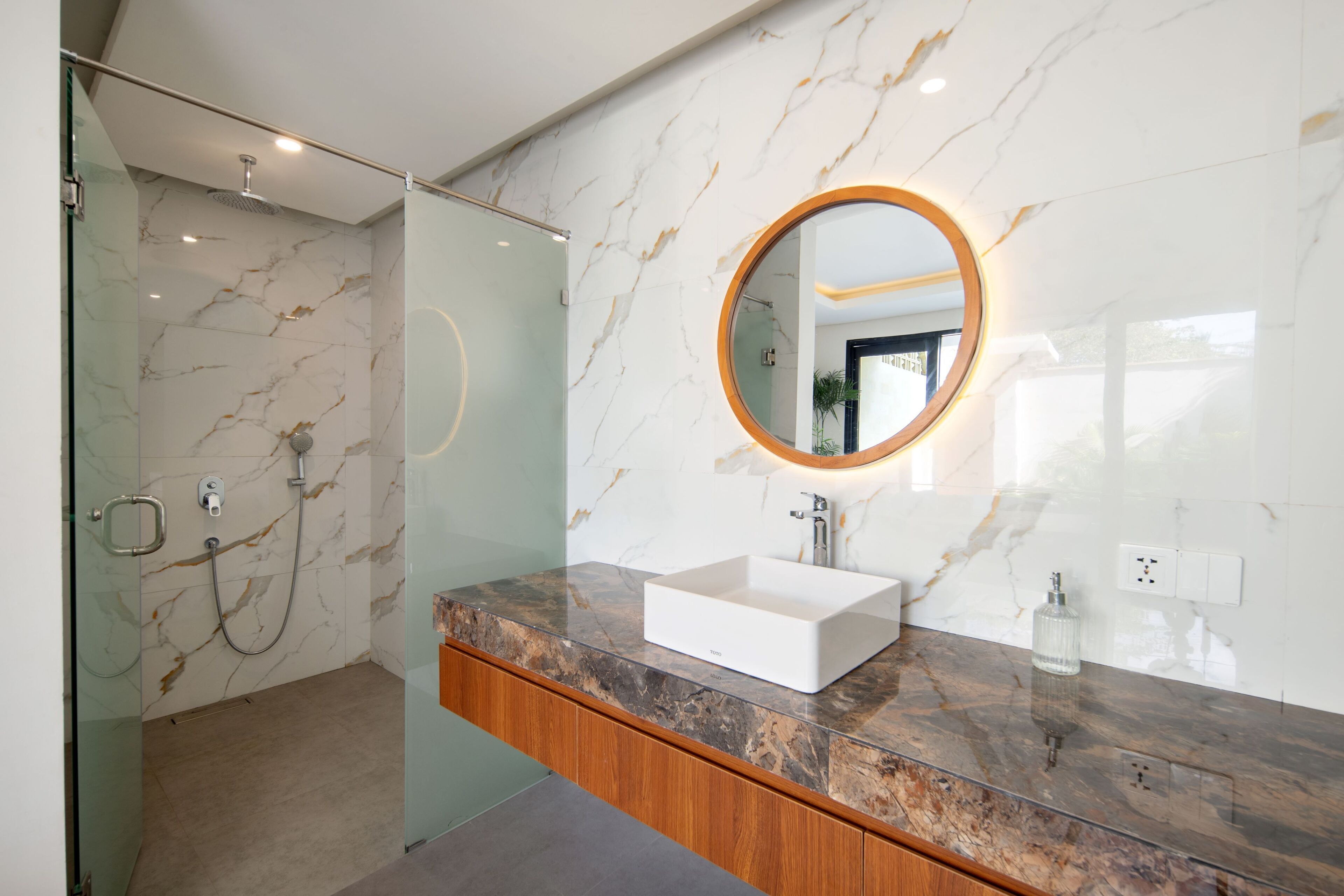 Un baño moderno con cabina de ducha de vidrio, pared con azulejos de mármol, tocador de madera y un espejo redondo con luz.