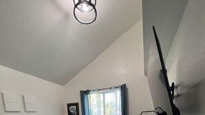 Une chambre à coucher avec un lit, une fenêtre avec des rideaux, un luminaire au plafond et un téléviseur fixé au mur.