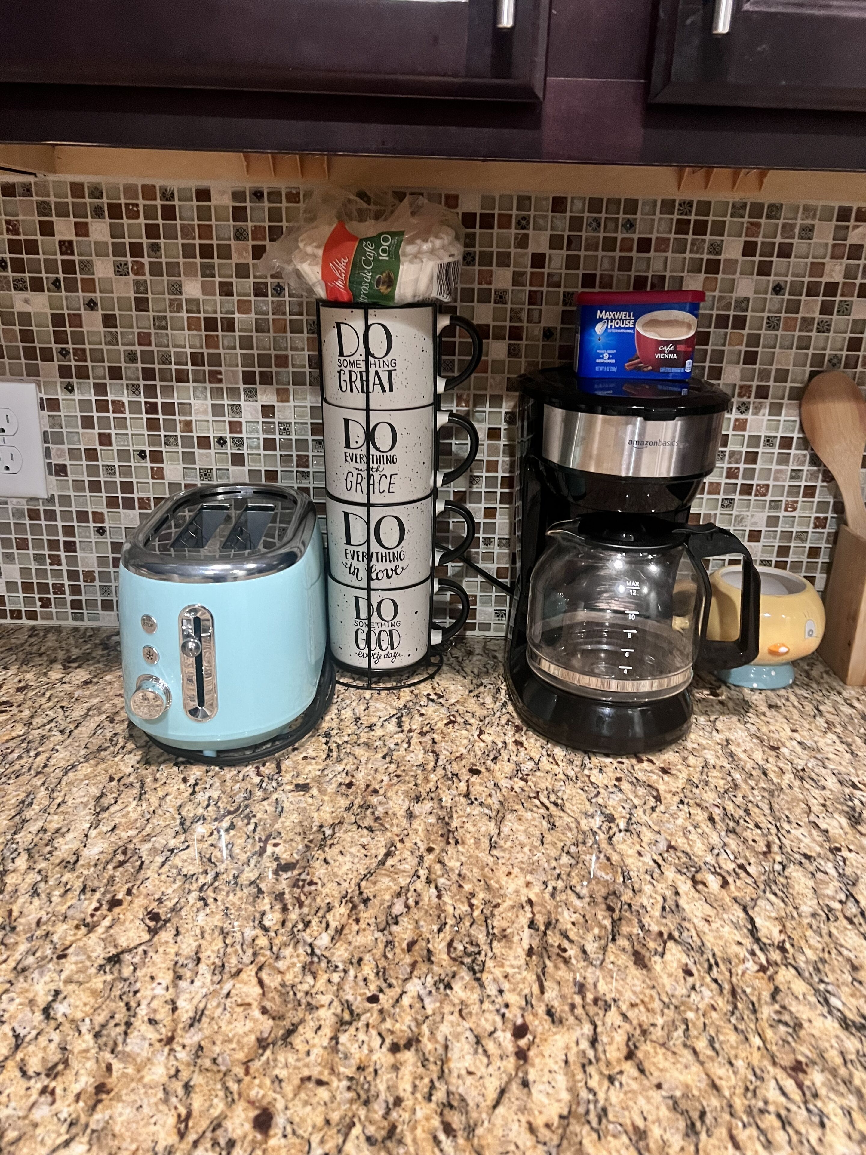 A kitchen counter with a toaster, a coffee maker, and a stack of mugs with motivational quotes.