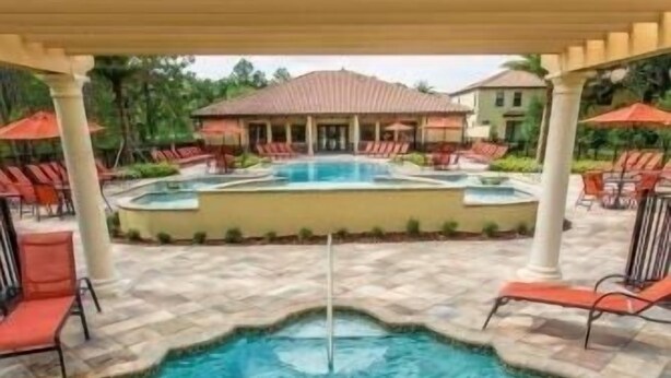 A swimming pool with a unique shape, surrounded by lounge chairs and umbrellas, under a covered area with a building in the background. - ChampionsGate Townhome + Fast WiFi, Beautiful Kitchen. Oasis Pool & Jacuzzi. (Davenport)