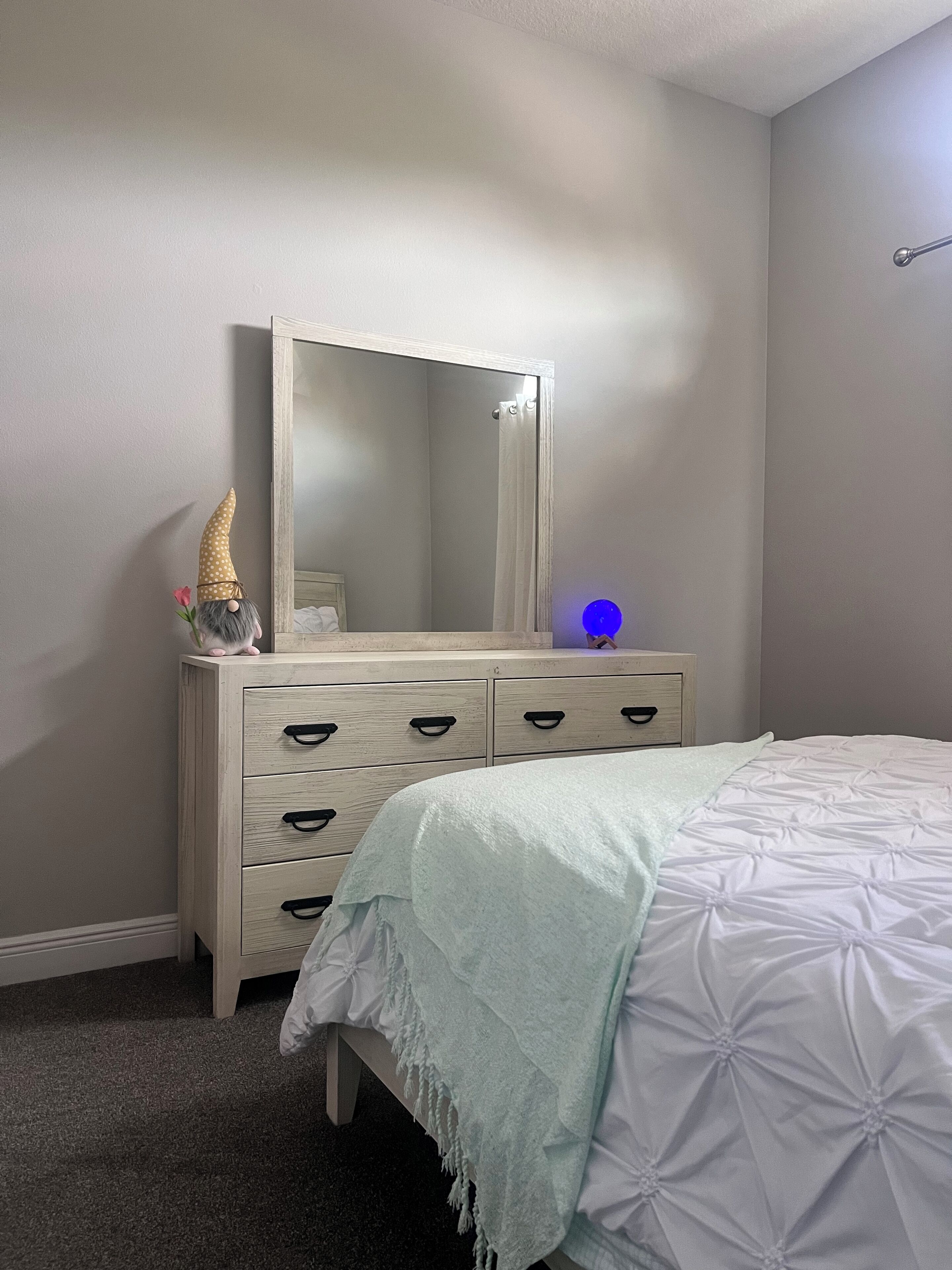 Une chambre à coucher comprenant un lit, une commode avec des tiroirs, un miroir et un luminaire fixé au mur.