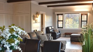 A modern living room with a dark wooden dining table, leather chairs, and a white vase with flowers. There is a sofa with cushions and a large window providing natural light. - Newly Renovated family-sized apartment downtown, lots of light!  (Storm Lake)