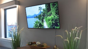 A modern living room with a flat-screen TV mounted on the wall, a wooden cabinet with decorative items, and two large potted plants. - Newly Renovated family-sized apartment downtown, lots of light!  (Storm Lake)