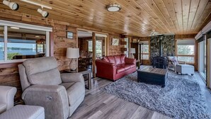 Een ruime woonkamer met houten wanden en plafond, een rode leren bank, een beige fauteuil en een stenen open haard.