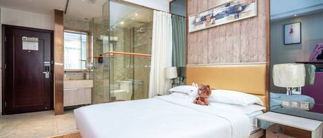 A hotel room with a large bed, a wooden headboard, a glass shower enclosure, a wooden floor, and a small bedside table with a lamp.